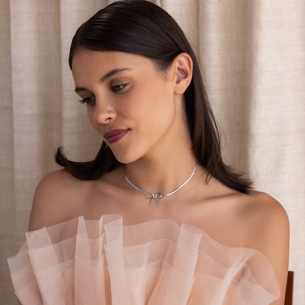 A brunette woman in an off-shoulder sheer dress wears the Herringbone Bow Necklace in Sterling Silver, gazing down thoughtfully with a coquette vibe.