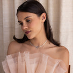A brunette woman in an off-shoulder sheer dress wears the Herringbone Bow Necklace in Sterling Silver, gazing down thoughtfully with a coquette vibe.