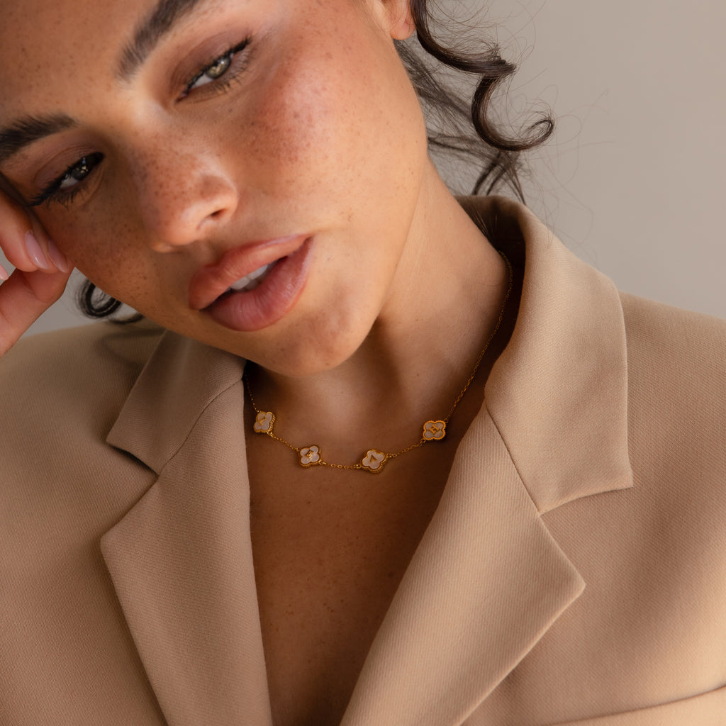 Woman in a beige blazer wears the Pearl Clover Letter Necklace, featuring gold flower-shaped pendants, as she looks toward the camera.