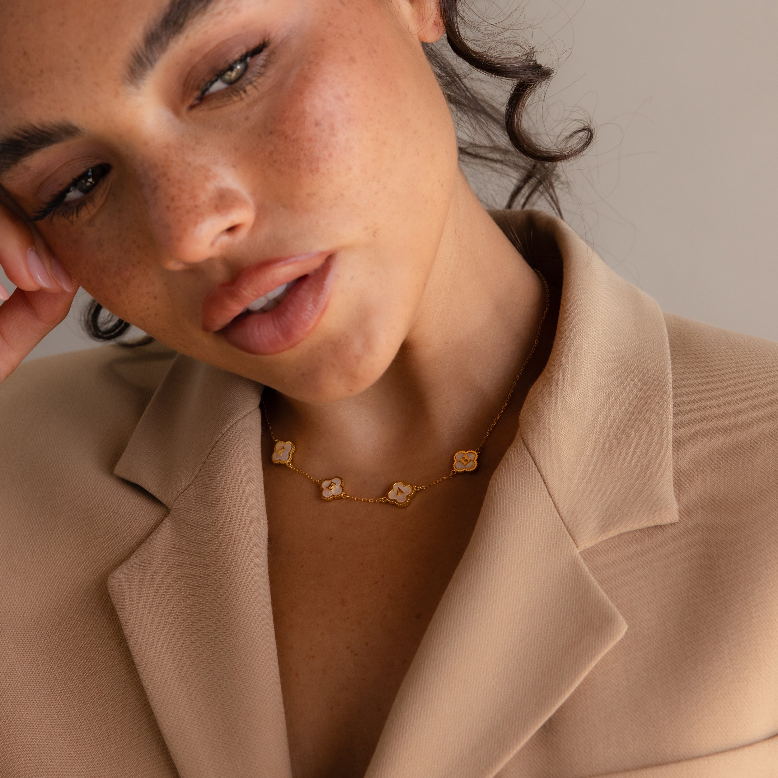 A person in a beige blazer wears the Gemstone Clover Letter Necklace, featuring four flower-shaped pendants, and poses with their head slightly tilted.