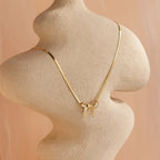 The Herringbone Bow Necklace in 18K Gold drapes elegantly on a curvy beige sculpture, highlighting its coquette charm against a soft peach background.