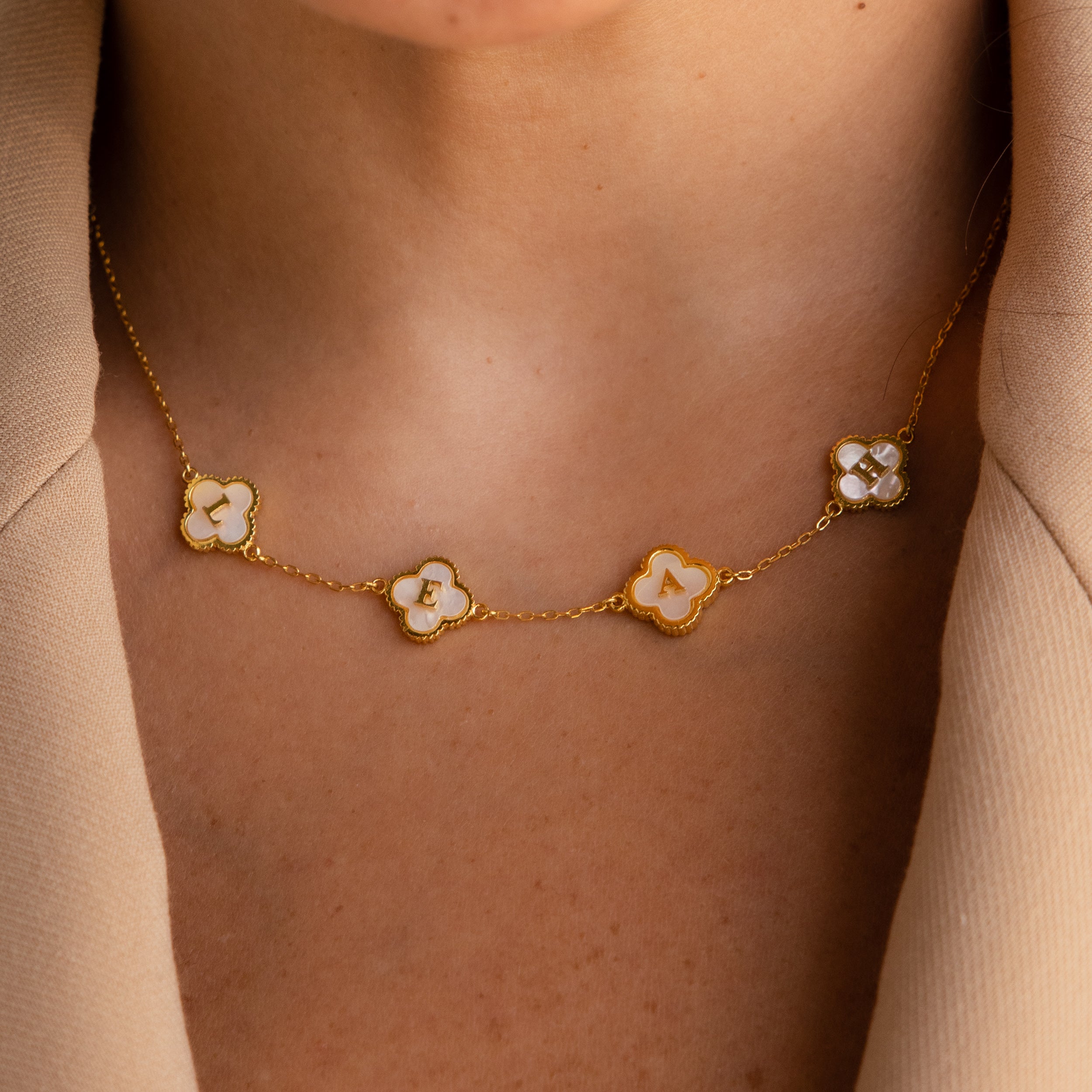 Close-up of the Pearl Clover Letter Necklace featuring white four-leaf clover charms, styled over a beige blazer.