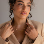 Woman with wavy hair in a beige blazer, wearing the Pearl Clover Letter Necklace with dainty gold chains, looking thoughtfully to the side.
