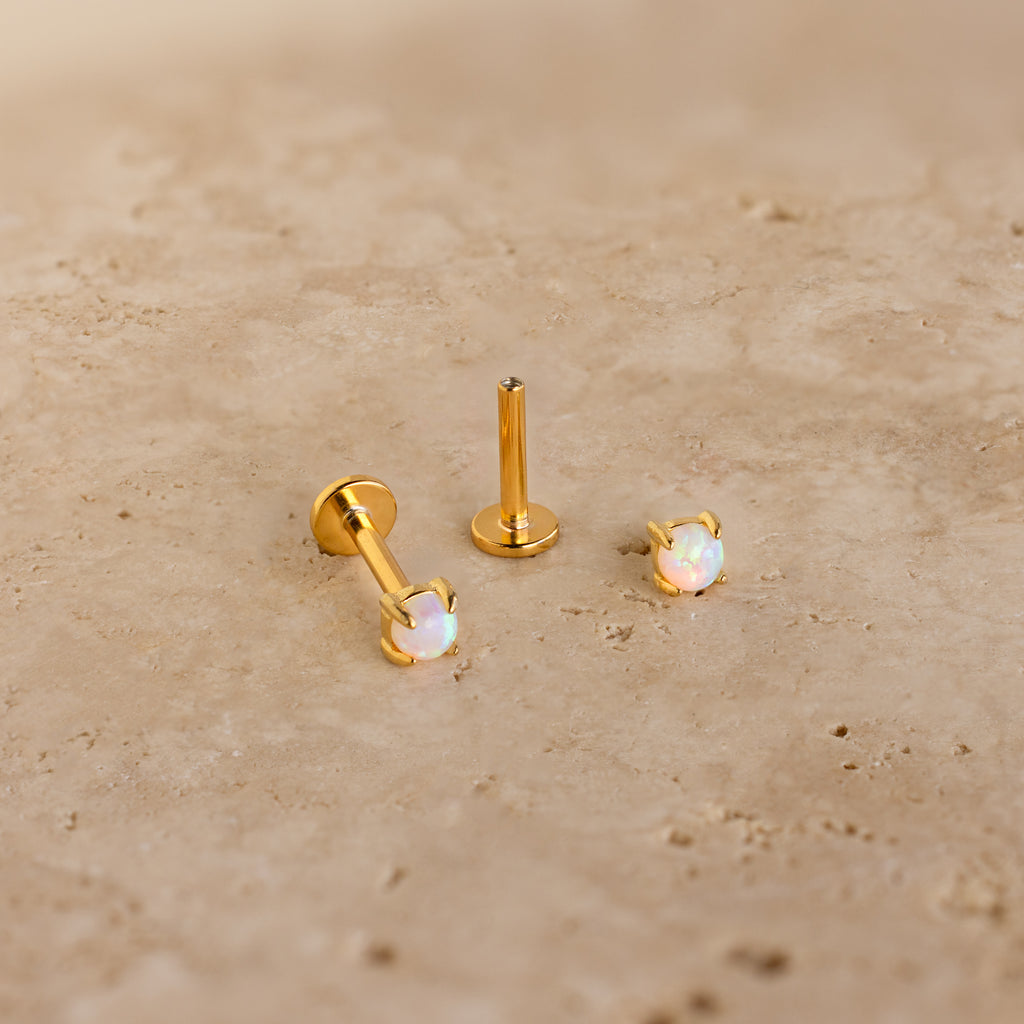 Two Opal Flat Back Studs in 18K Gold, adorned with iridescent opals, rest on a beige stone surface—ideal for anyone seeking dainty earrings with subtle elegance.