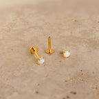 Two Opal Flat Back Studs in 18K Gold, adorned with iridescent opals, rest on a beige stone surface—ideal for anyone seeking dainty earrings with subtle elegance.