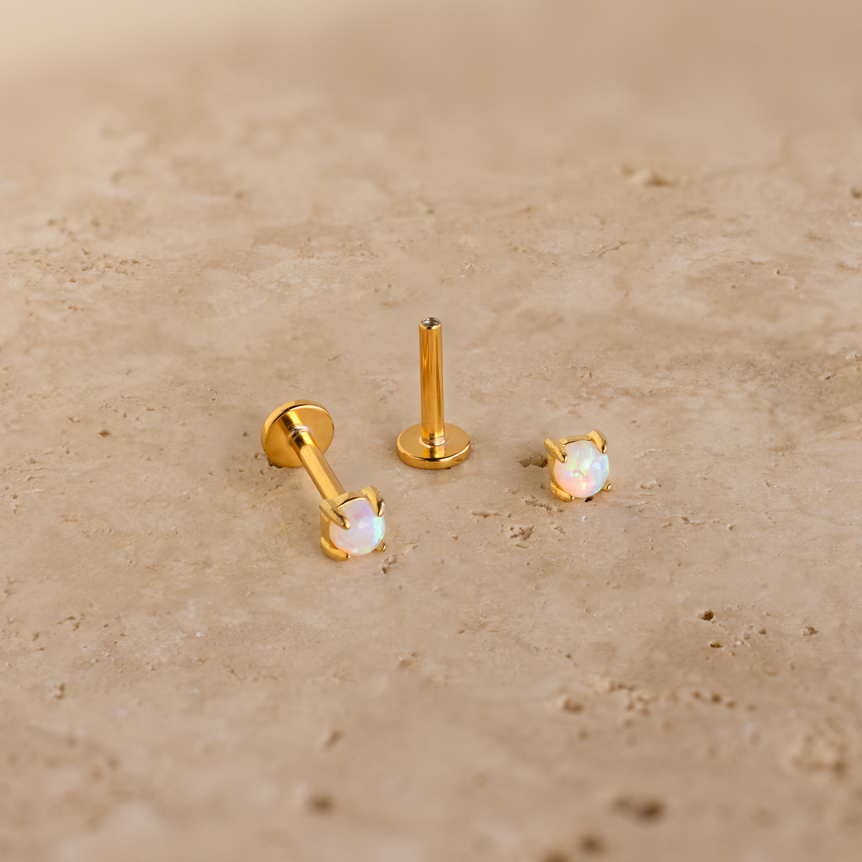 Two Opal Flat Back Studs in 18K Gold, adorned with iridescent opals, rest on a beige stone surface—ideal for anyone seeking dainty earrings with subtle elegance.