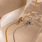 Gold and silver ribbons tied in bows on beige fabric evoke coquette style, resembling the layered look of the Herringbone Bow Necklace, with a sheer ribbon mimicking elegant necklace layers in the background.