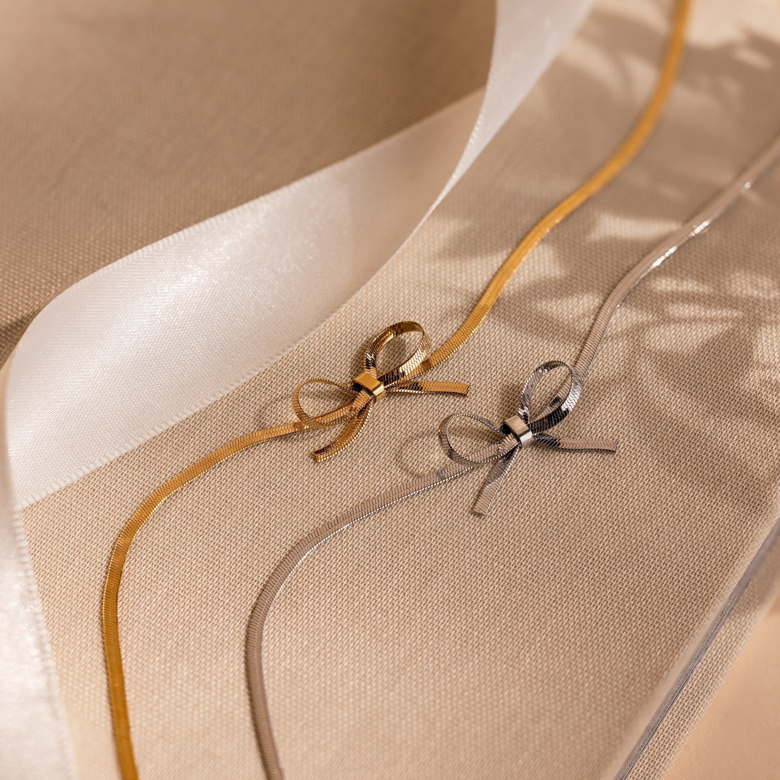 Gold and silver ribbons tied in bows on beige fabric evoke coquette style, resembling the layered look of the Herringbone Bow Necklace, with a sheer ribbon mimicking elegant necklace layers in the background.