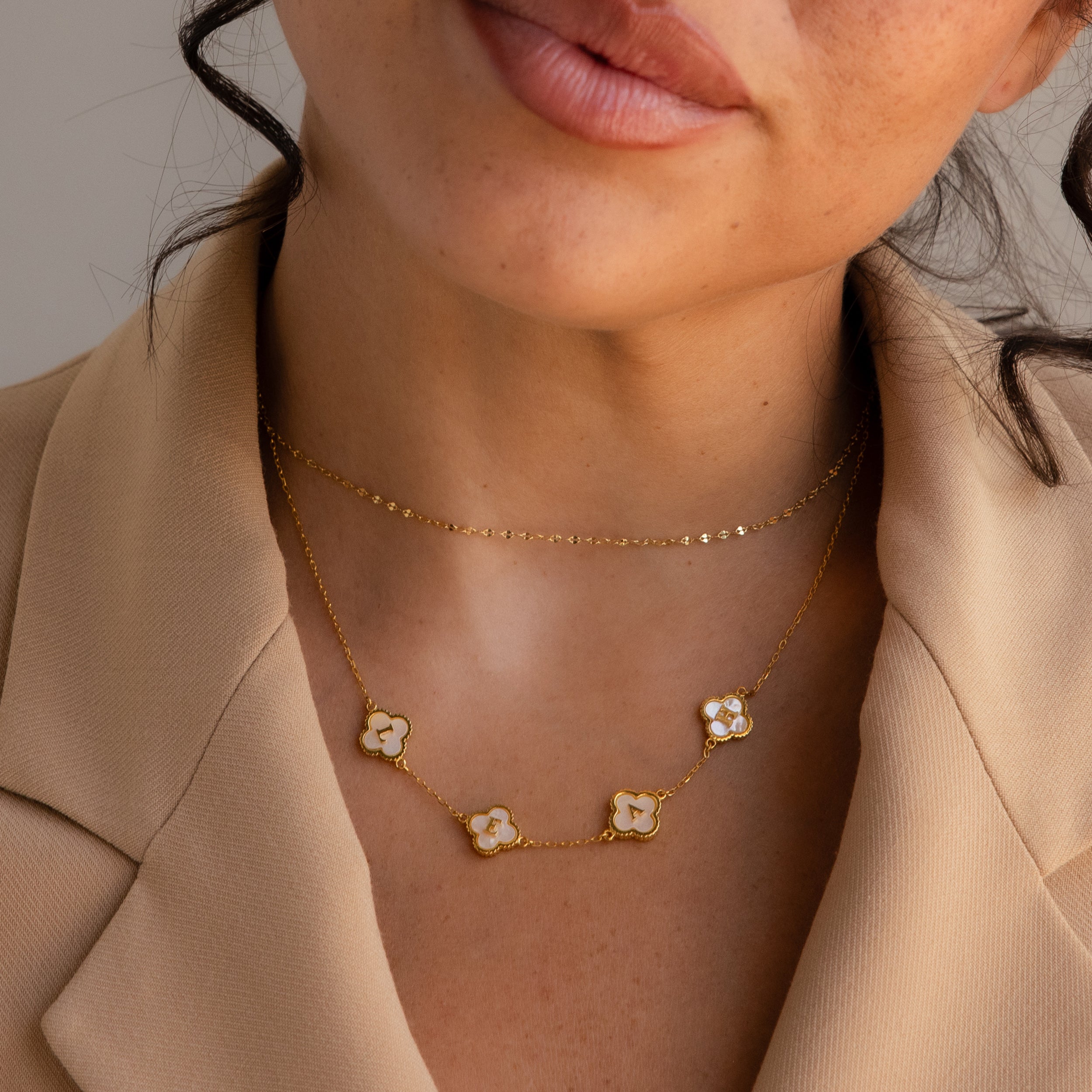 A woman pairs a beige blazer with layered gold necklaces, including the Pearl Clover Letter Necklace and flower pendants.