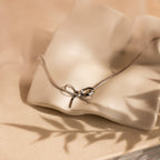 The Herringbone Bow Necklace in Sterling Silver rests on a wavy, cream ceramic dish—perfect for adding a touch of coquette style to any look.