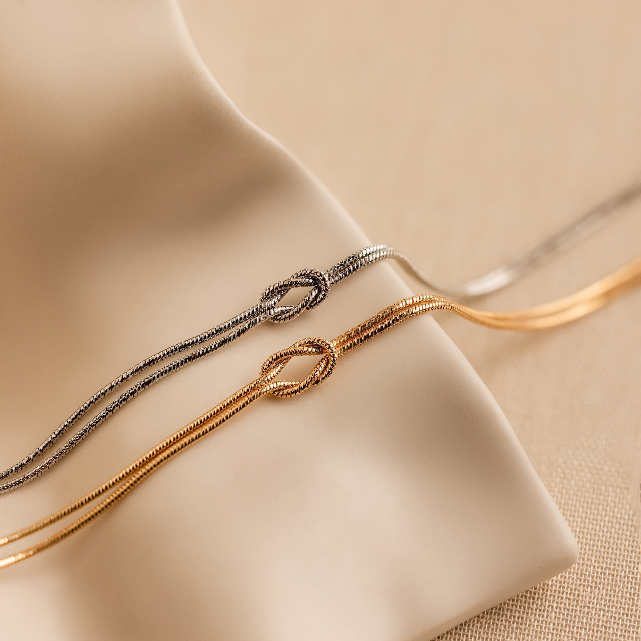 Two elegant Love Knot Bracelets—one gold, one silver—are displayed on soft beige fabric, making them the perfect gift for couples.