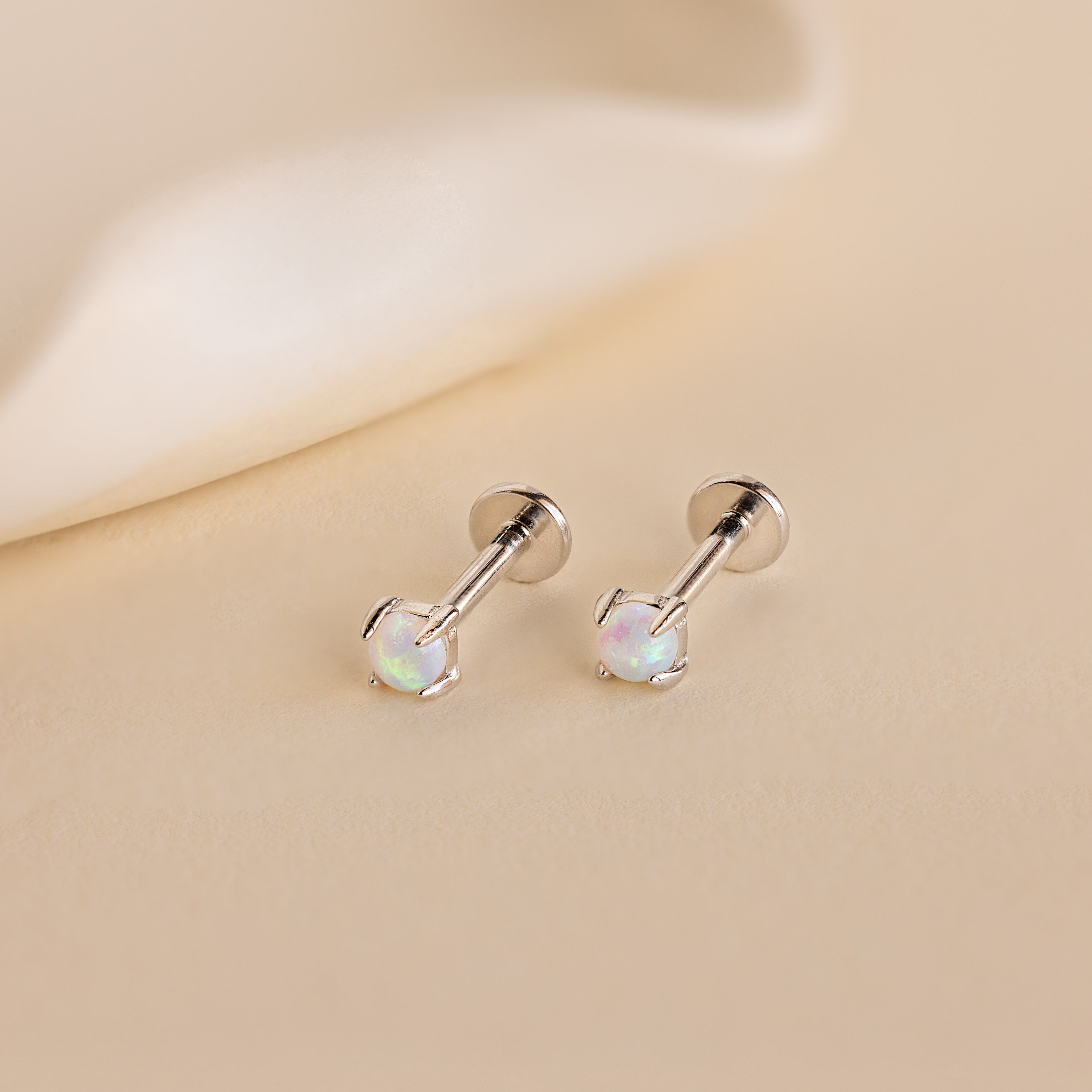 The Opal Flat Back Studs in Sterling Silver are comfortable sleeper earrings featuring round opal gemstones, displayed as silver studs on a light beige surface.