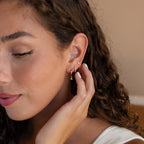 A woman with curly hair touches her ear, highlighting the Classic Diamond Earrings Set in 18K Gold—timeless wedding earrings or elegant diamond gemstone accents for any occasion, complemented by a natural makeup look.