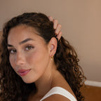 A woman with curly hair in a white top smiles softly and touches her hair while showcasing the Classic Diamond Earrings Set in 18K Gold.