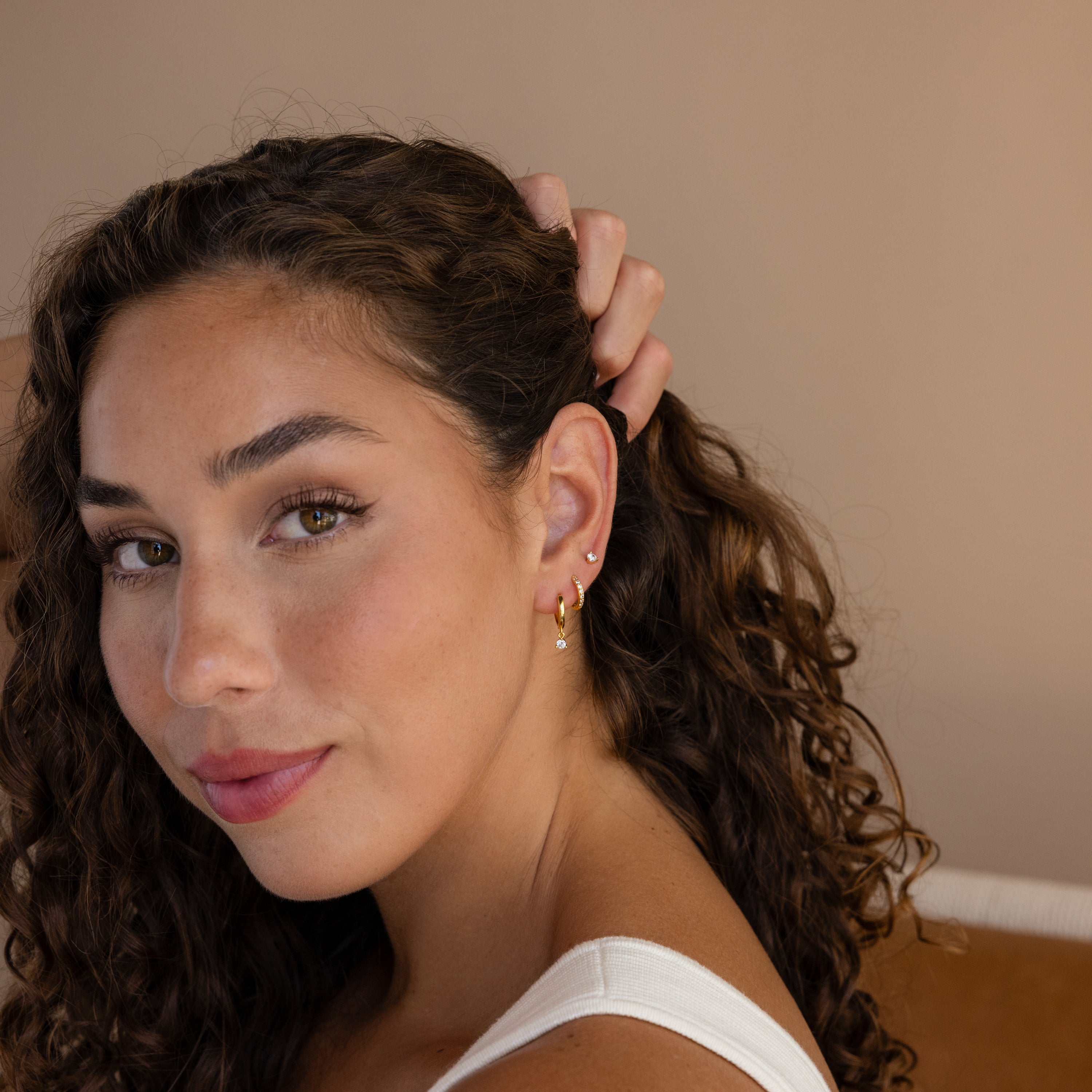 A woman with curly hair in a white top smiles softly and touches her hair while showcasing the Classic Diamond Earrings Set in 18K Gold.