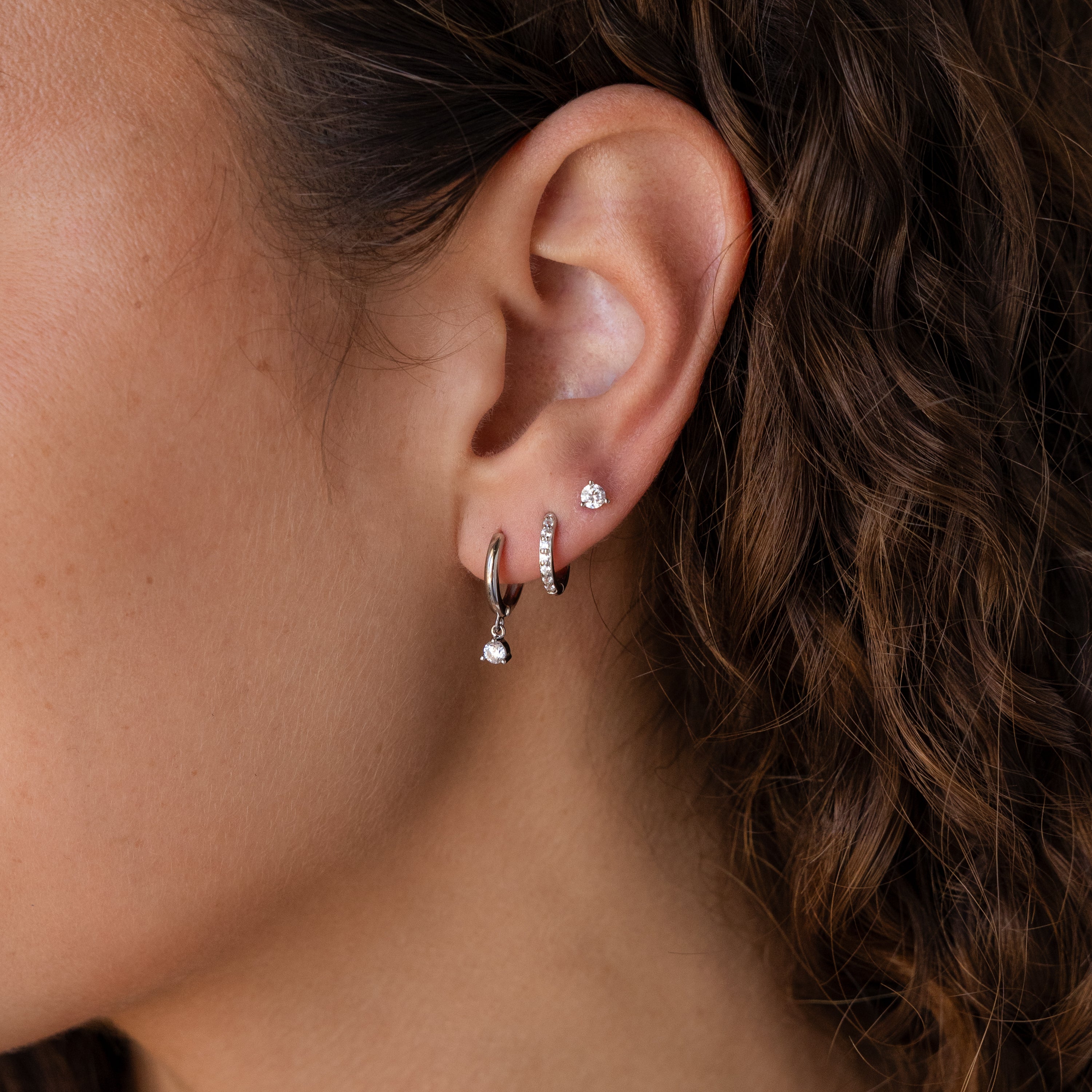 Close-up of an ear with three silver hoops and a classic stud from the Classic Diamond Earrings Set, next to curly brown hair.