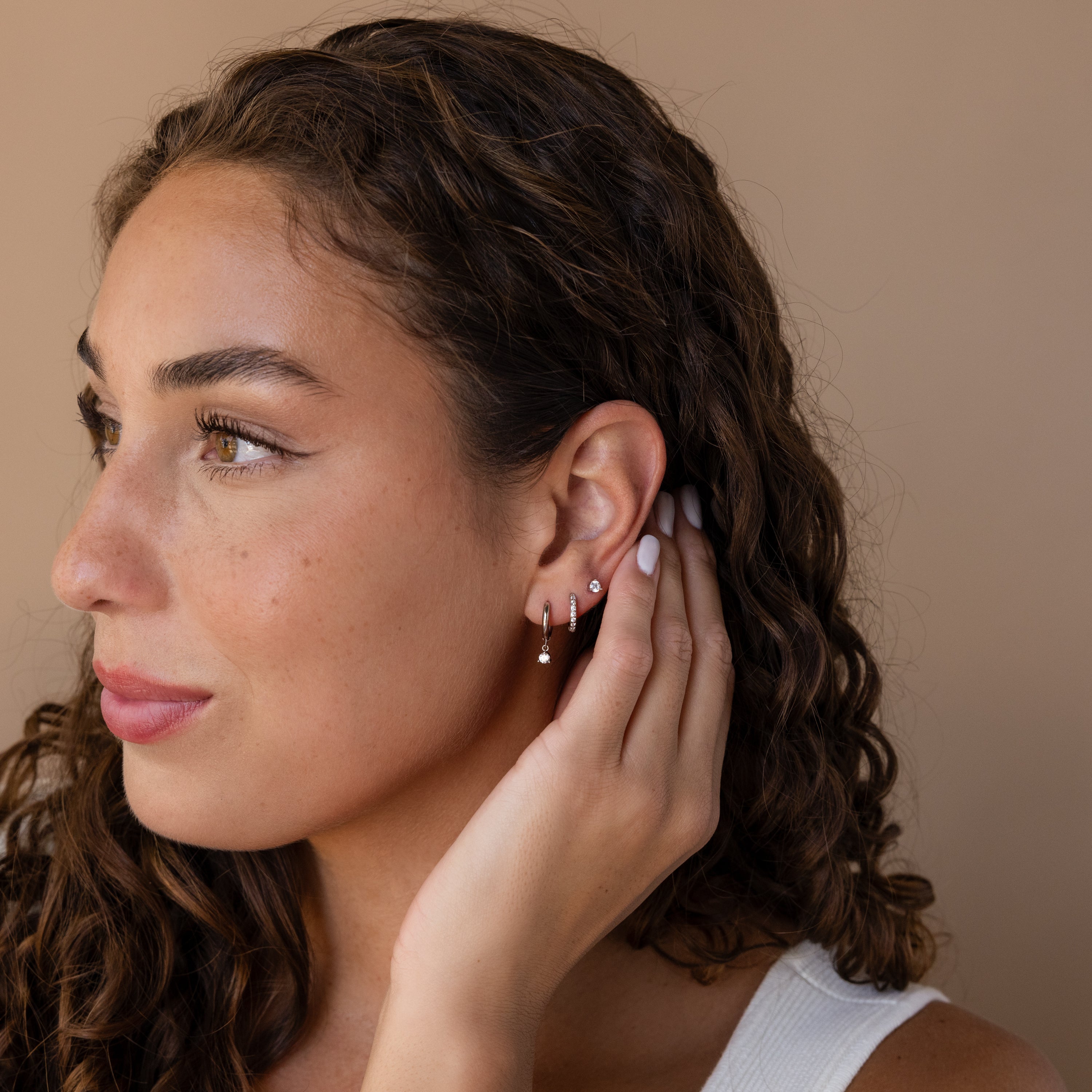 A woman with curly hair touches her ear while wearing the Classic Diamond Earrings Set in Sterling Silver, featuring multiple small hoops and a classic stud, as she looks to the left.