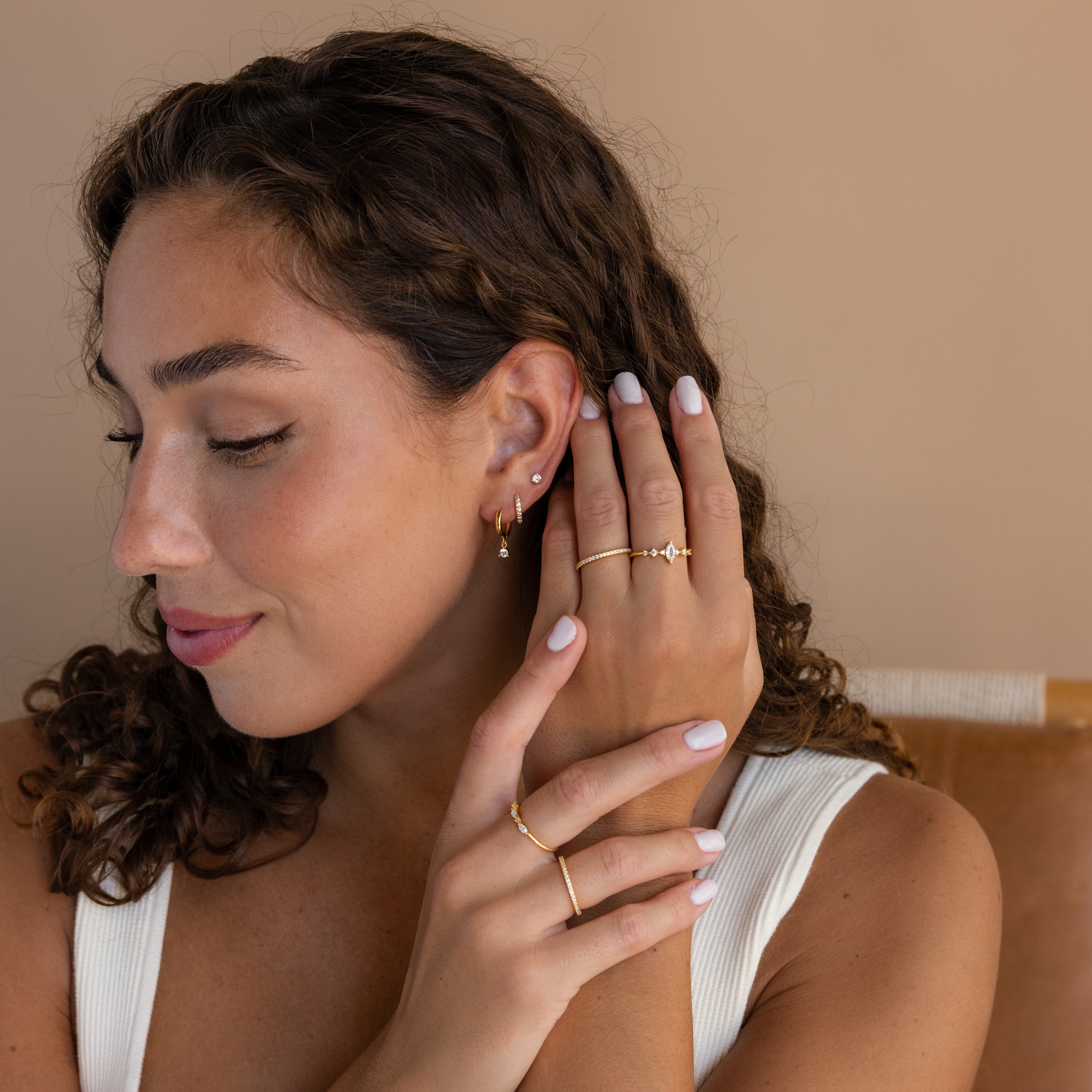 Woman with curly hair wears Classic Diamond Earrings Set in 18K Gold and gold rings, looking down with her hand near her face against a neutral background.