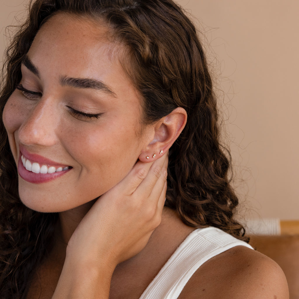A smiling woman with curly hair touches her ear, wearing a white top and showcasing the Tiny Diamond Flatback Studs Set for a subtle touch of sparkle.