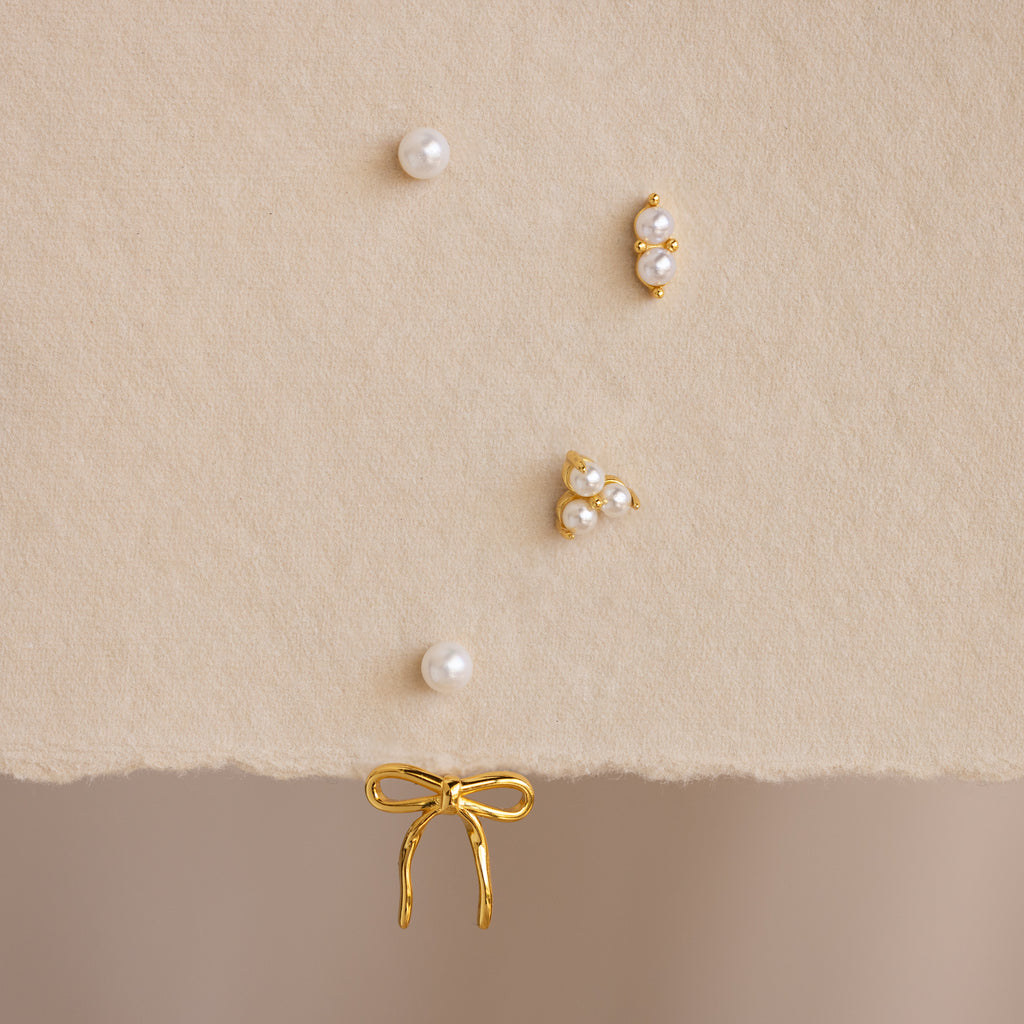 The Pearl & Bow Ear Jackets in 18K Gold, featuring five assorted gold and pearl studs including elegant bow designs, are displayed on a soft beige textured surface.
