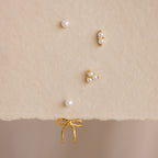 The Pearl & Bow Ear Jackets in 18K Gold, featuring five assorted gold and pearl studs including elegant bow designs, are displayed on a soft beige textured surface.