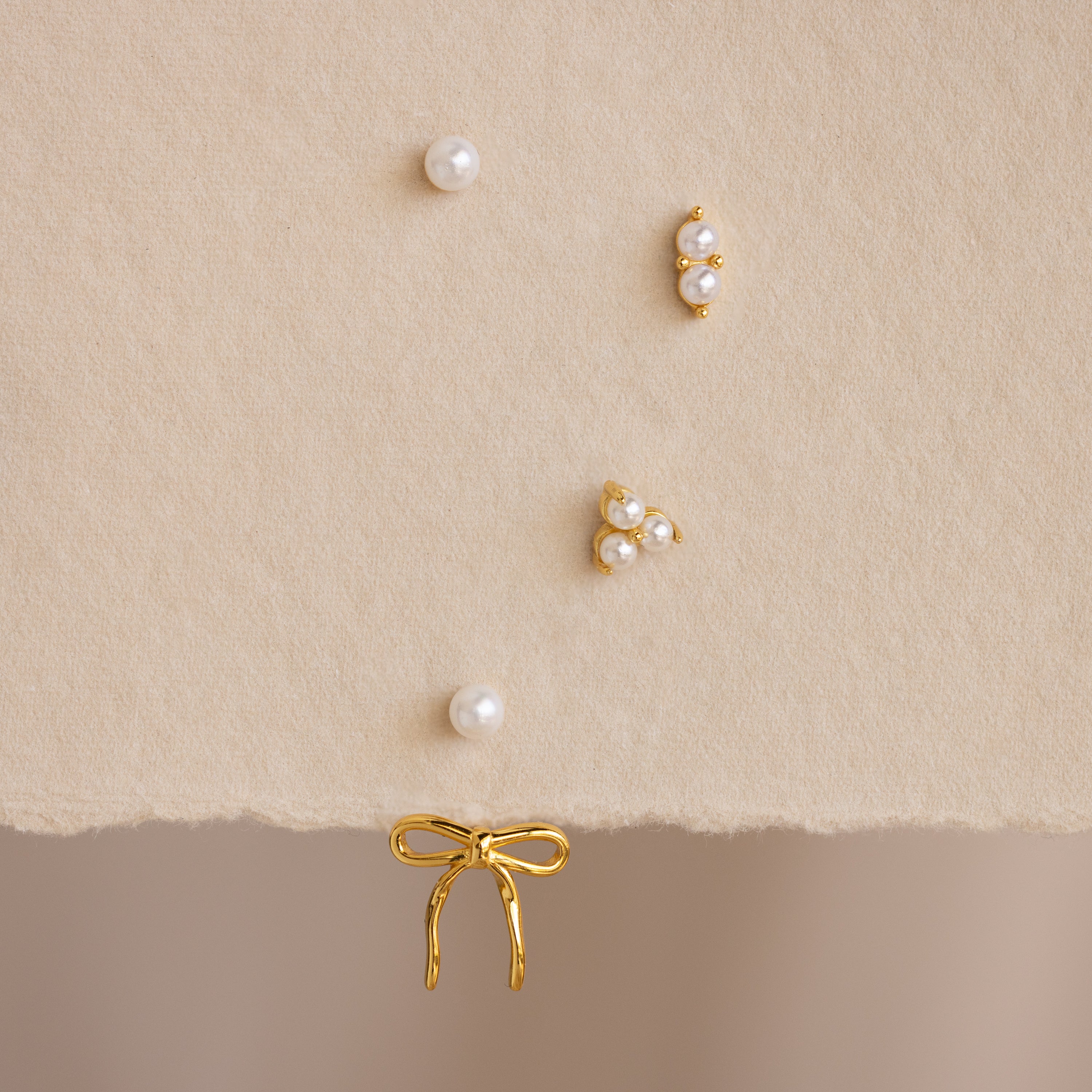 The Pearl & Bow Ear Jackets in 18K Gold, featuring five assorted gold and pearl studs including elegant bow designs, are displayed on a soft beige textured surface.
