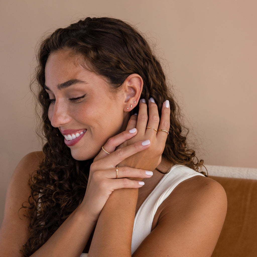 Woman with curly hair smiles, wearing gold rings and dainty earrings, including the Tiny Diamond Flatback Studs Set in 18K Gold, as her hands gently touch her face and neck.