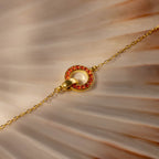 The Interlocking Birthstone Bracelet features a gold chain and a red gemstone-studded circular charm, shown on a seashell background. Personalize this elegant bracelet to add a unique touch to your jewelry collection.