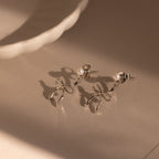 The Pearl & Bow Ear Jackets in Sterling Silver feature delicate pearl accents in a unique bow design, resting elegantly on a neutral surface near a white ceramic dish.