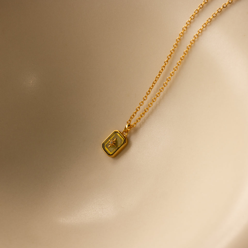 The North Star Birthstone Necklace in Peridot (August) features a rectangular pendant with a star motif, reminiscent of a diamond-set North Star, shown on a smooth beige surface.
