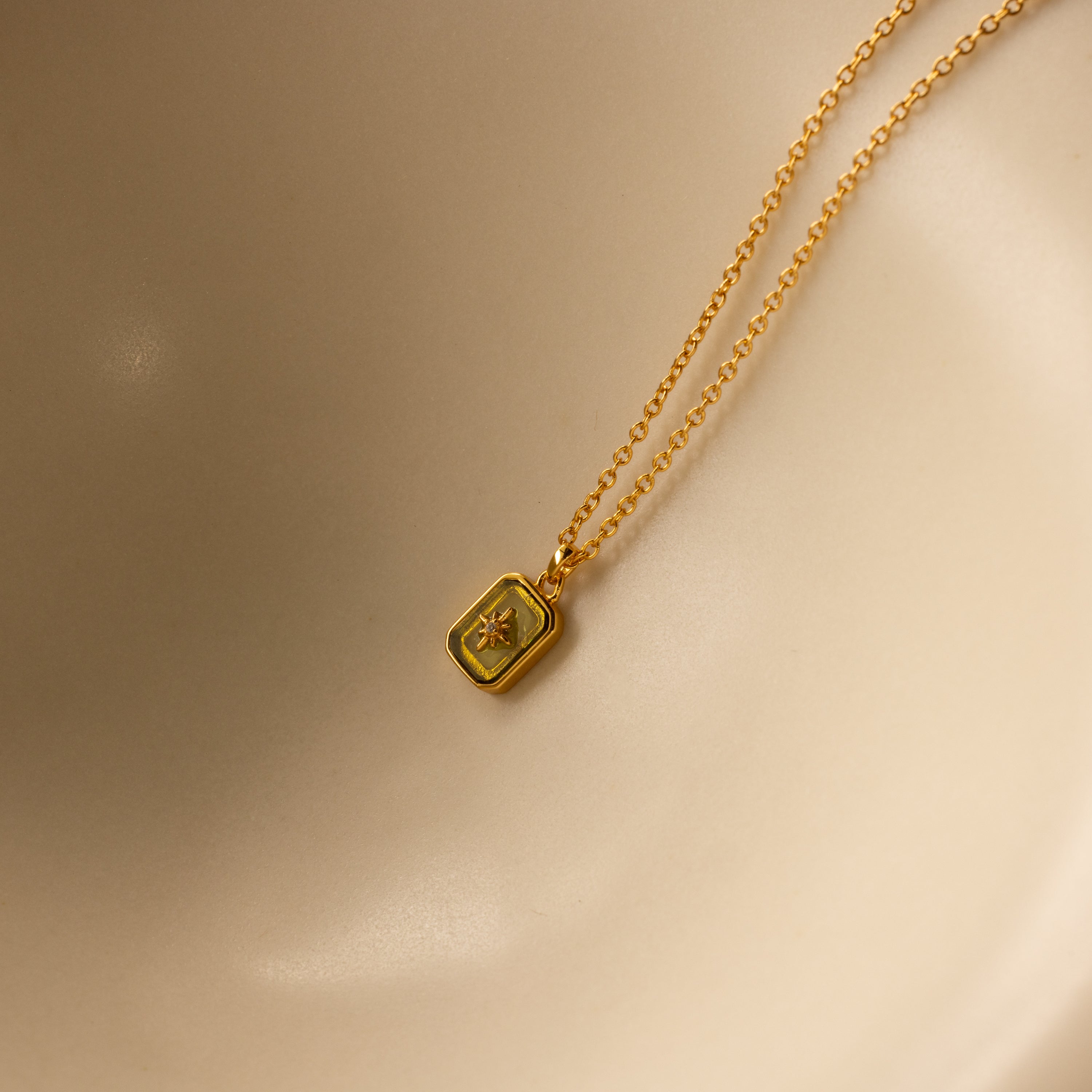 The North Star Birthstone Necklace in Peridot (August) features a rectangular pendant with a star motif, reminiscent of a diamond-set North Star, shown on a smooth beige surface.
