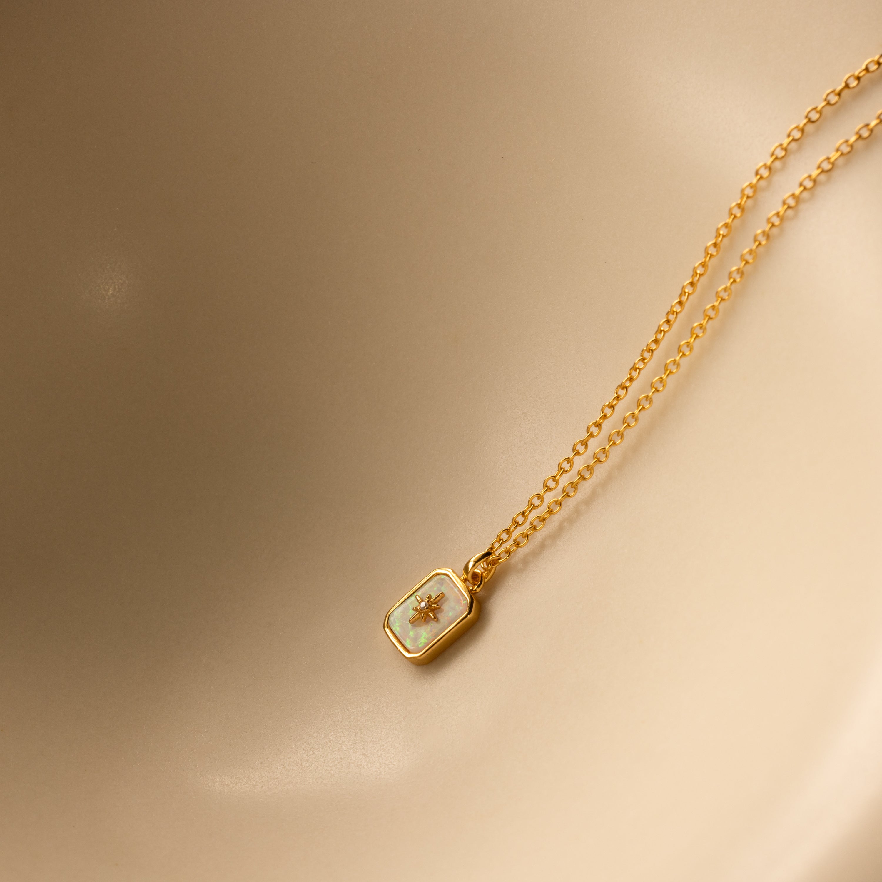 North Star Opal Necklace: A gold chain necklace with a rectangular pendant featuring a small star design and opal detail, displayed on a beige background.