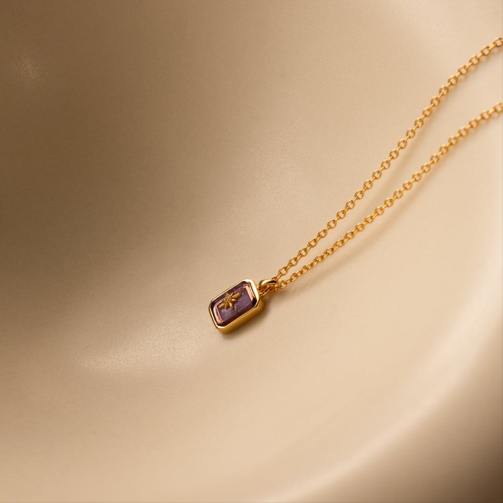 North Star Birthstone Necklace in Amethyst (February) featuring a rectangular purple crystal pendant on a gold chain, displayed on a smooth beige background.