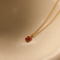 The North Star Birthstone Necklace in Ruby (July) features a small red rectangular crystal pendant with a diamond-set North Star detail, displayed on a beige surface.