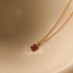 The North Star Birthstone Necklace in Ruby (July) features a small red rectangular crystal pendant with a diamond-set North Star detail, displayed on a beige surface.