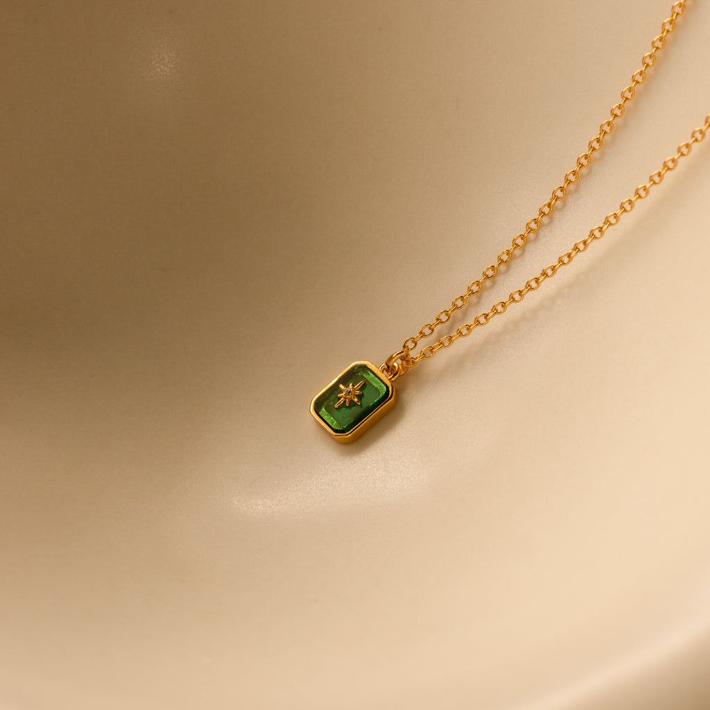 North Star Birthstone Necklace in Emerald (May) with a rectangular crystal pendant, displayed on a beige curved surface.