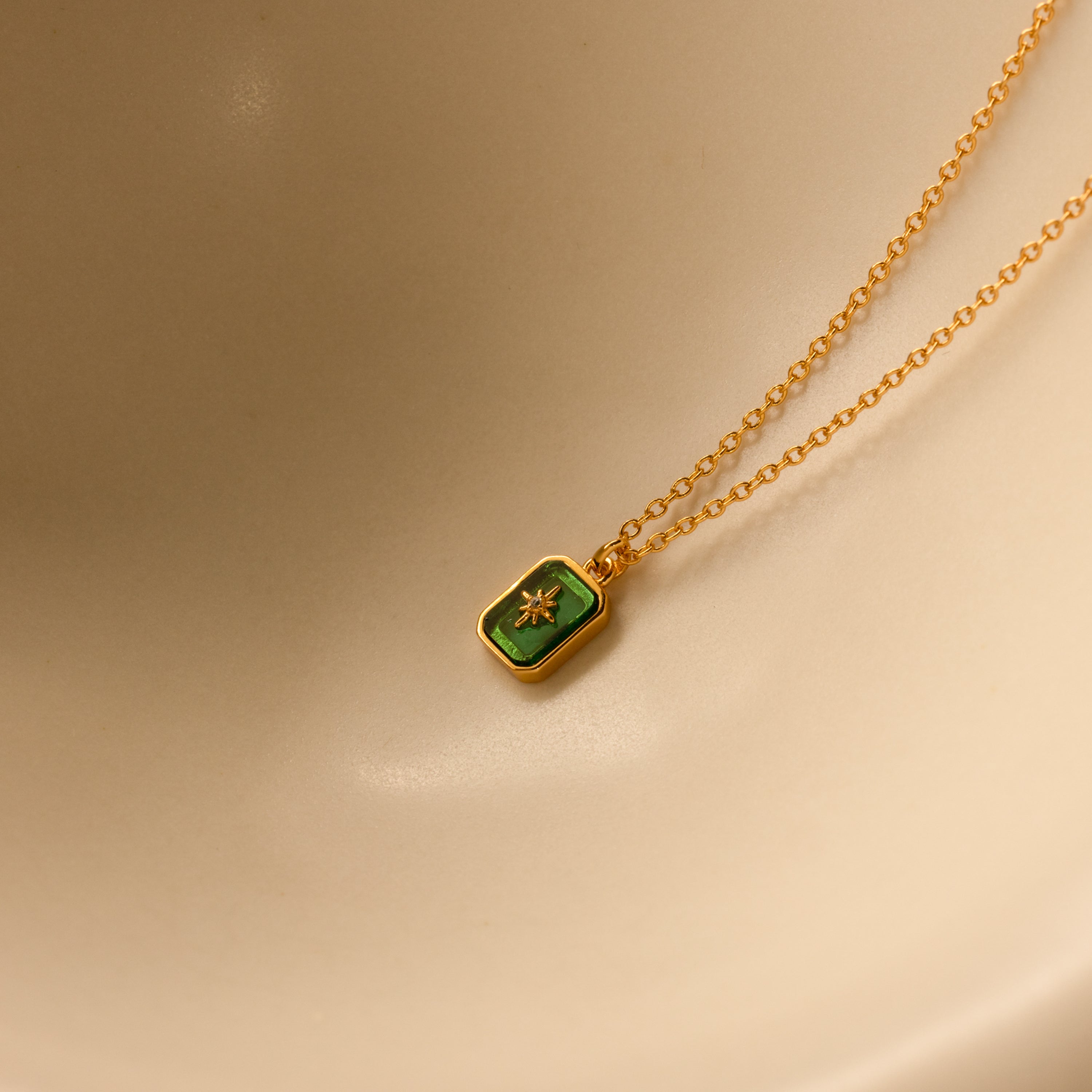 North Star Birthstone Necklace in Emerald (May) with a rectangular crystal pendant, displayed on a beige curved surface.