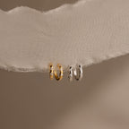 The Mini Diamond Sprinkle Huggies—one gold, one silver—dangle on ivory fabric against a beige background, offering subtle sparkle in a perfect petite hoop style.