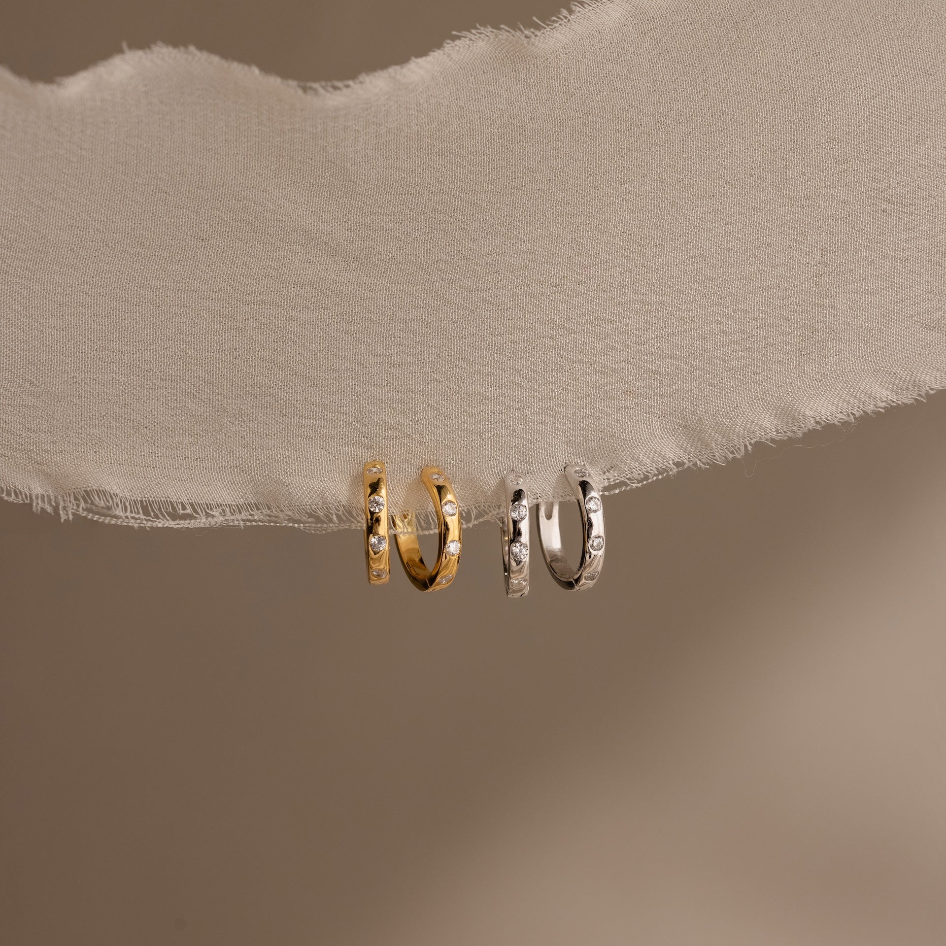 The Mini Diamond Sprinkle Huggies—one gold, one silver—dangle on ivory fabric against a beige background, offering subtle sparkle in a perfect petite hoop style.