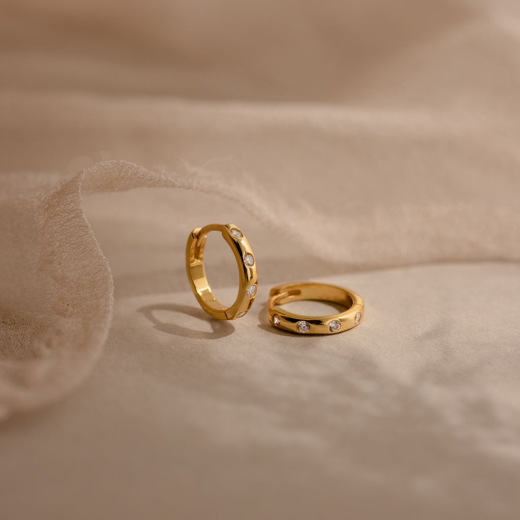 Two gold rings with small diamonds rest on soft beige fabric, styled alongside Mini Diamond Sprinkle Huggies in 18K Gold for a timeless, elegant look.