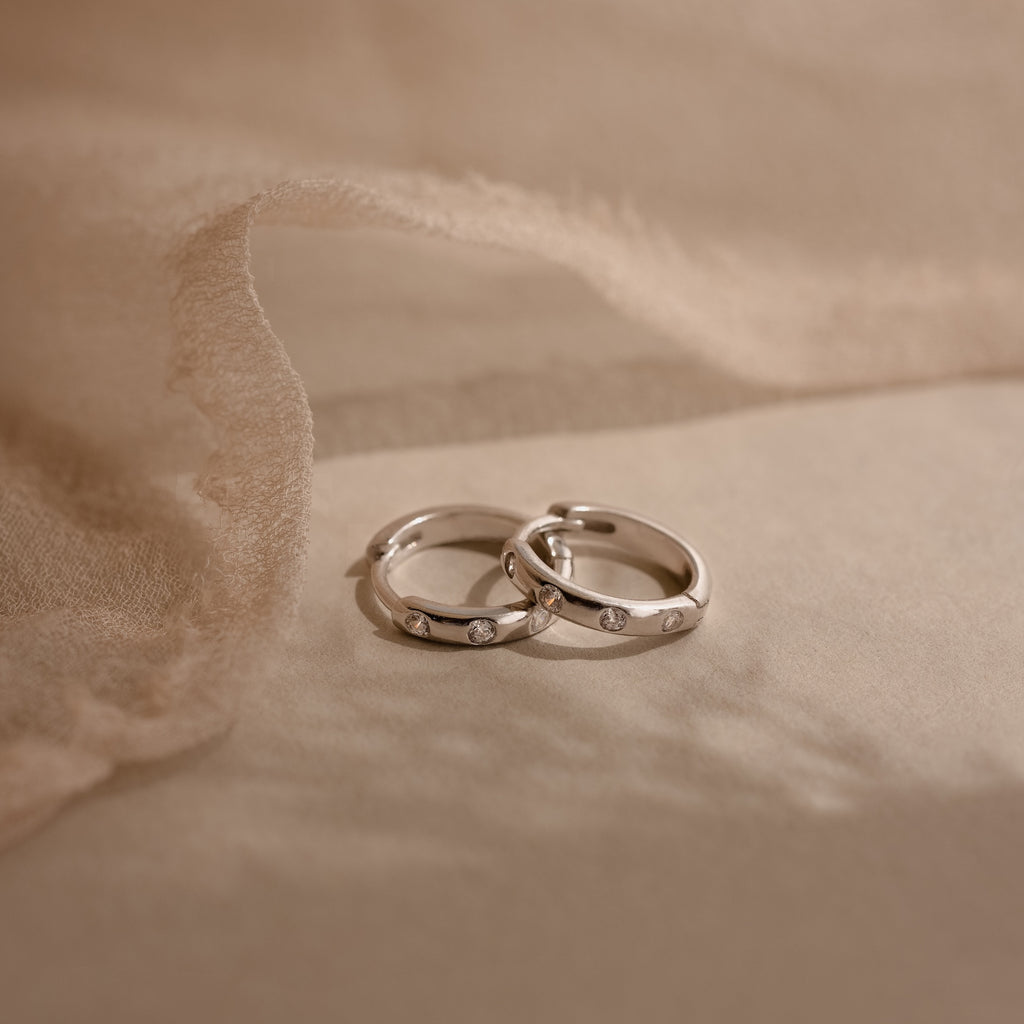 Two Mini Diamond Sprinkle Huggies in Silver rest on a soft beige fabric background, their sparkling diamonds evoking the elegance of delicate silver hoops.