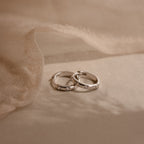 Two Mini Diamond Sprinkle Huggies in Silver rest on a soft beige fabric background, their sparkling diamonds evoking the elegance of delicate silver hoops.