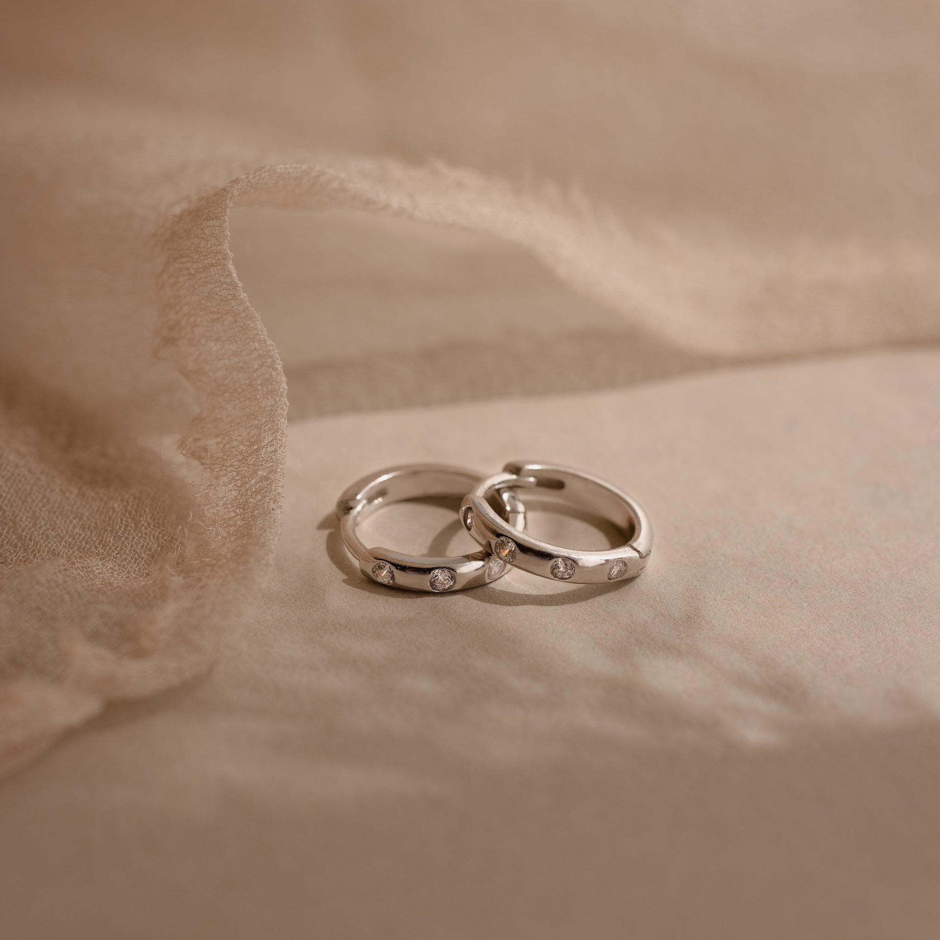 Two Mini Diamond Sprinkle Huggies in Silver rest on a soft beige fabric background, their sparkling diamonds evoking the elegance of delicate silver hoops.