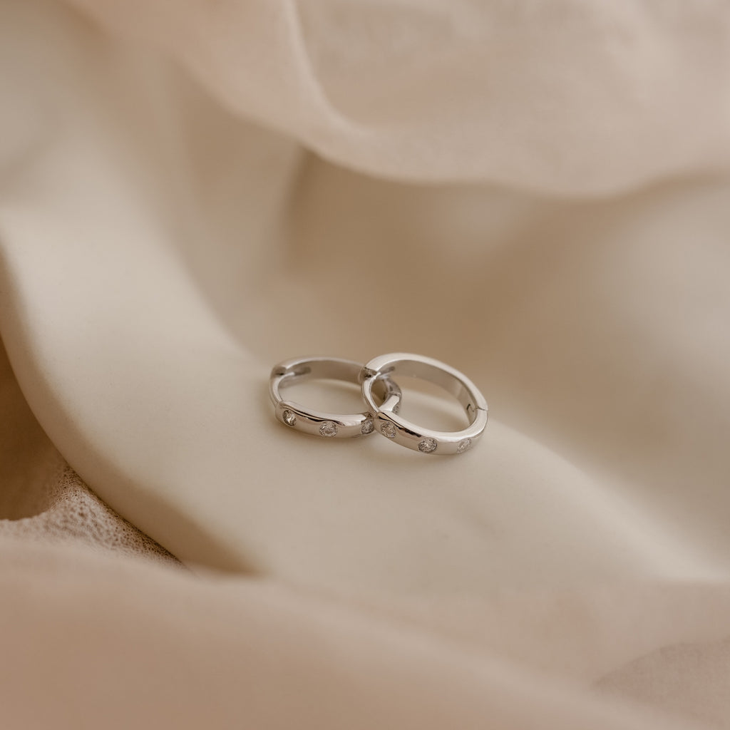 The Mini Diamond Sprinkle Huggies in Silver rest on soft beige fabric, their delicate diamonds sparkling gently.