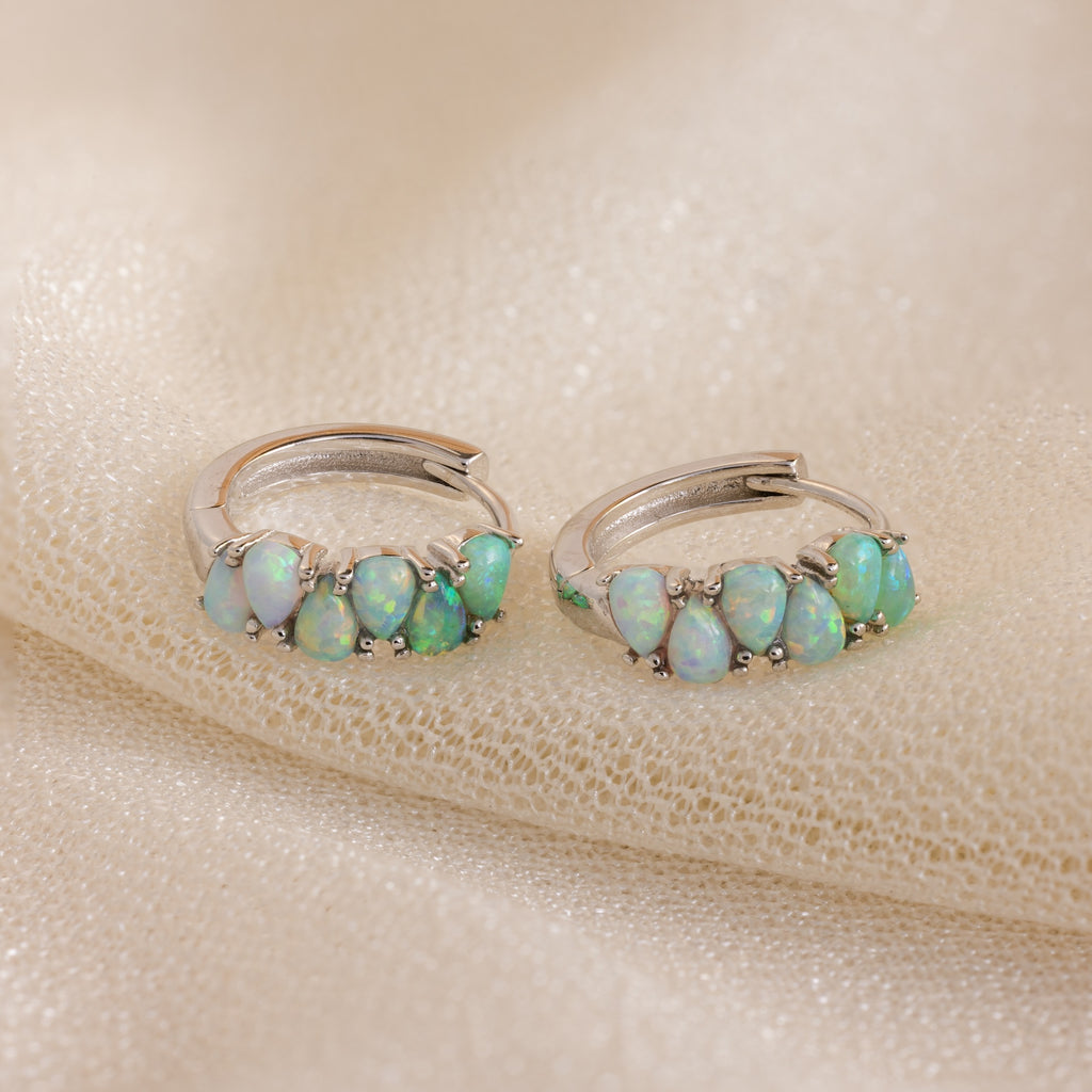 Ombre Opal Huggies featuring small oval opals on silver hoops, displayed on shimmery beige fabric.