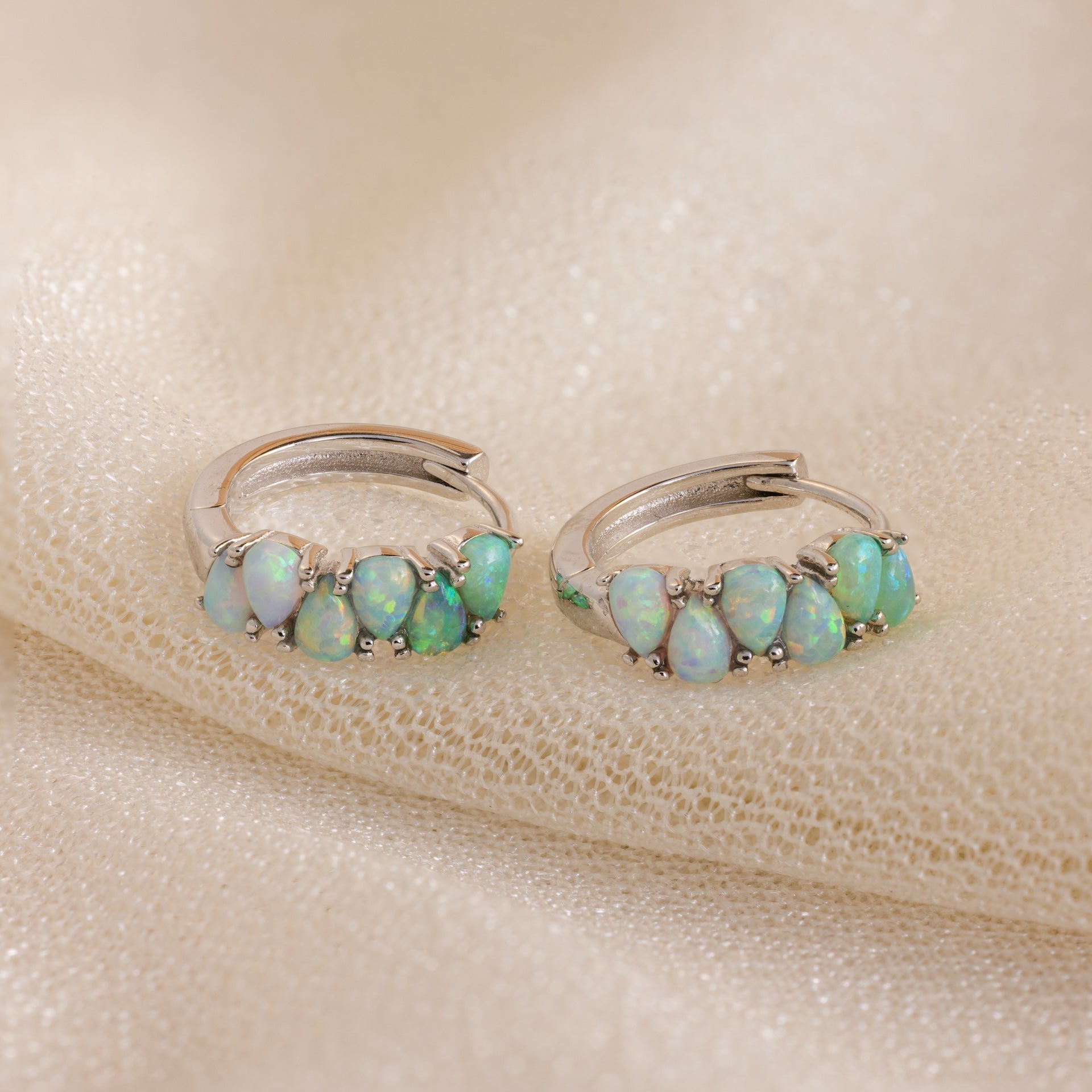 Ombre Opal Huggies featuring small oval opals on silver hoops, displayed on shimmery beige fabric.