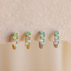 Four gold and silver rings with opals are displayed on a textured beige surface, shown alongside Ombre Opal Huggies and teardrop gemstones.