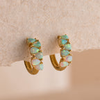 Ombre Opal Huggies in 18K Gold, featuring five oval opals each, are showcased on a textured beige surface.