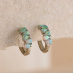 Ombre Opal Huggies in Silver with iridescent opal stones set in a row, displayed against a textured beige surface.
