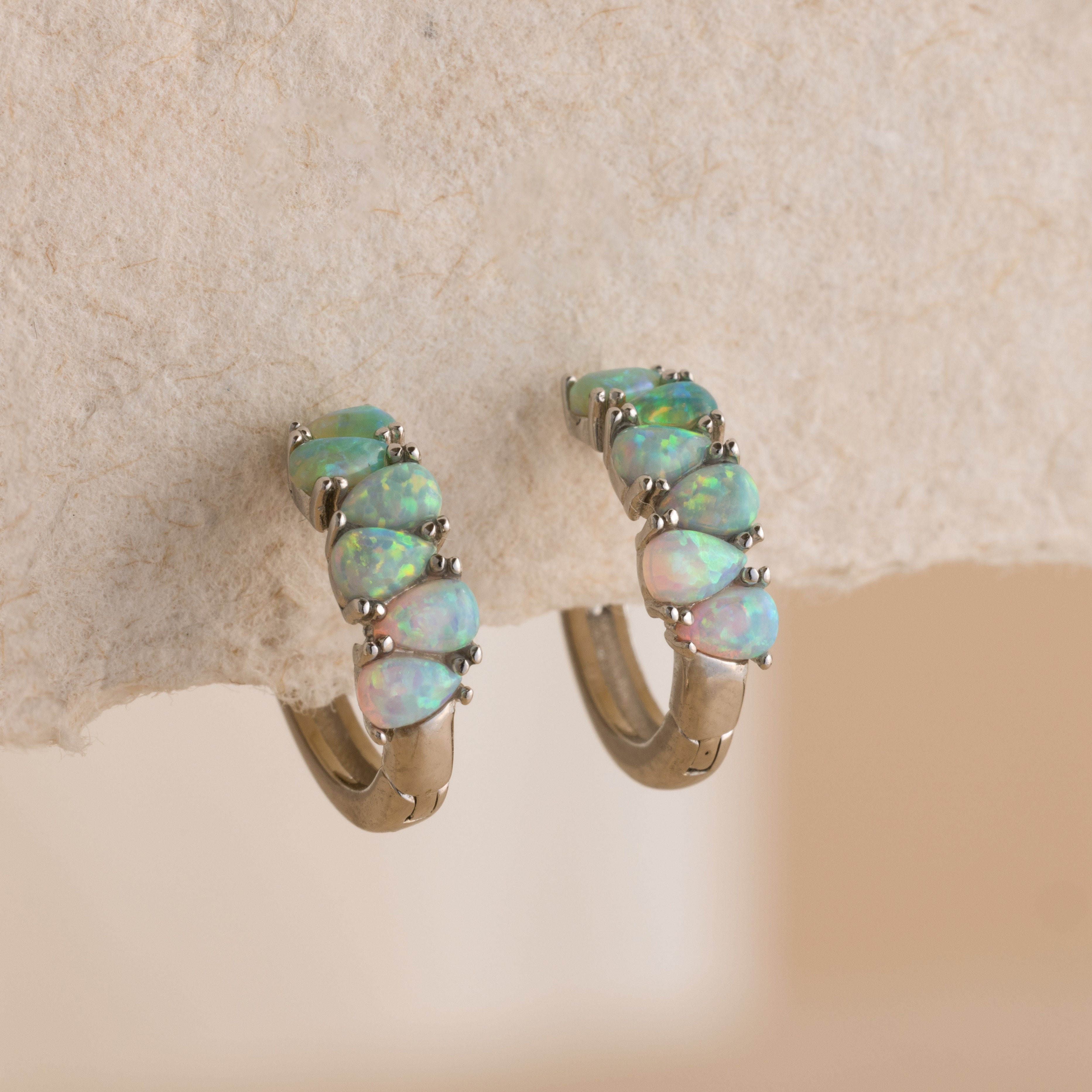 Ombre Opal Huggies in Silver with iridescent opal stones set in a row, displayed against a textured beige surface.